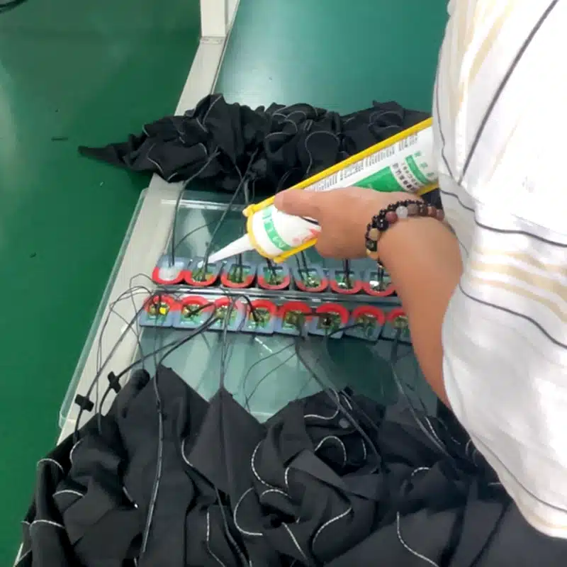Factory worker assembling heating elements for custom heated gloves, applying adhesive to secure heating pads for durable electric heated gloves production