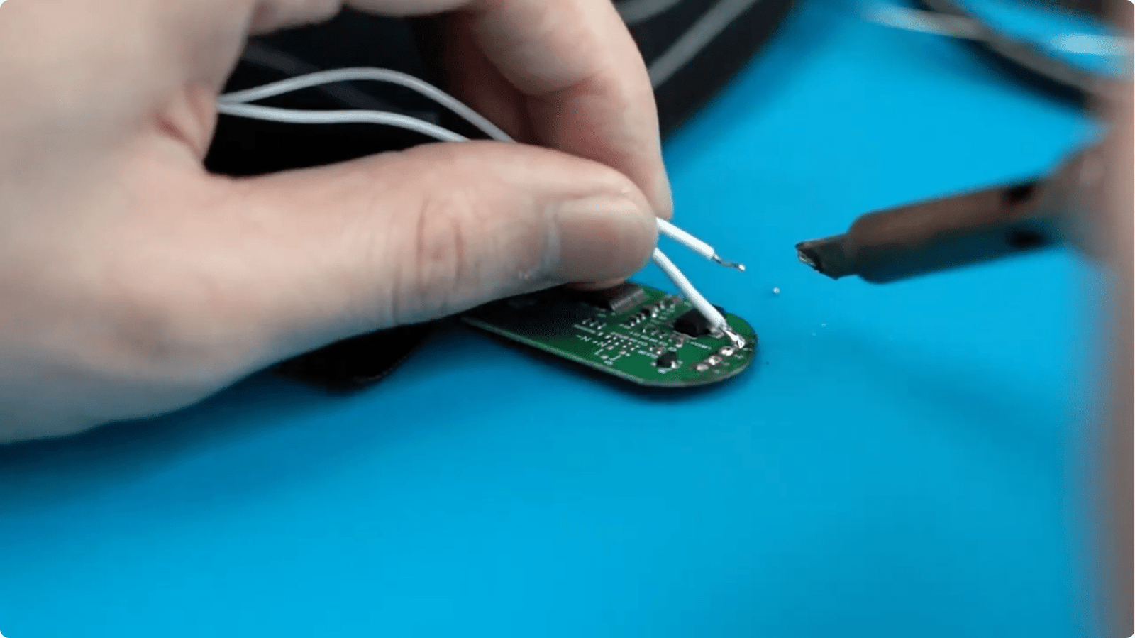 Close-up of technician hand-soldering white wires onto a green PCBA circuit board on blue ESD mat, precision assembly of heating control modules