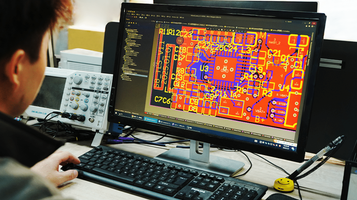 Engineer working on PCB circuit board layout design using EDA software, with red and yellow traces visible on screen and oscilloscope on desk