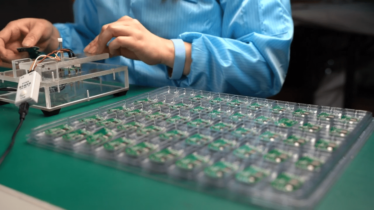 Technician in blue ESD smock testing PCBA heating control modules using test fixture, with organized trays of green circuit boards on anti-static mat