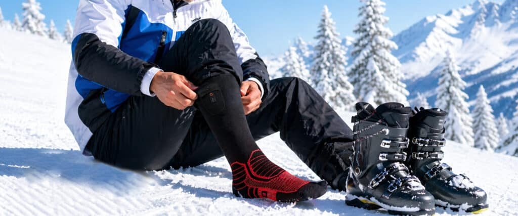 Skier putting on red and black heated socks on snowy mountain slope, ski boots beside them with snow-covered pine trees and alpine peaks in background