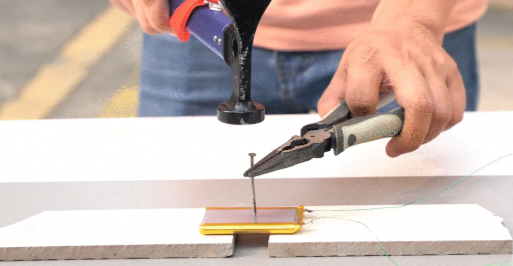 A technician uses pliers to hold a metal nail against a lithium-ion pouch cell, while a hammer is positioned to drive the nail through the battery, demonstrating a nail penetration safety test.